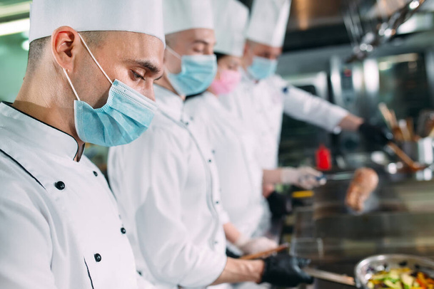 Chefs con máscaras protectoras y guantes preparan comida en la cocina de un restaurante u hotel. - Foto, Imagen
