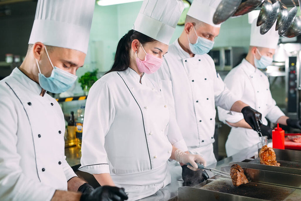 Chefs in protective masks and gloves prepare food in the kitchen of a restaurant or hotel. - Zdjęcie, obraz