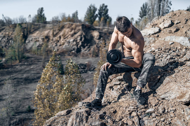 Full length photo of a bodybuilder with metal dumbbell in hand. Shirtless bodybuilder. Rocks on the background. - Фото, изображение