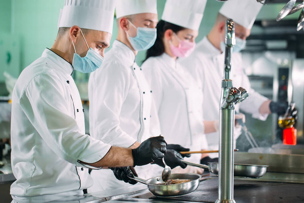 Chefs in protective masks and gloves prepare food in the kitchen of a restaurant or hotel. - 写真・画像