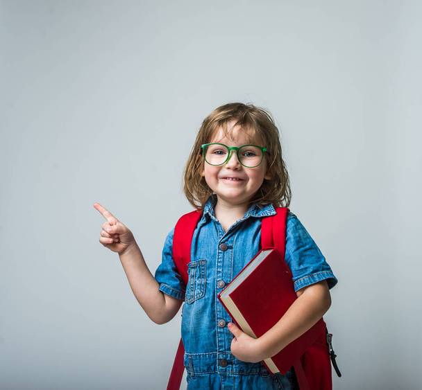 Funny portrait of happy little girl in glasses hugging a book. Child with backpack laughing and pointing with finger up. - Фото, зображення