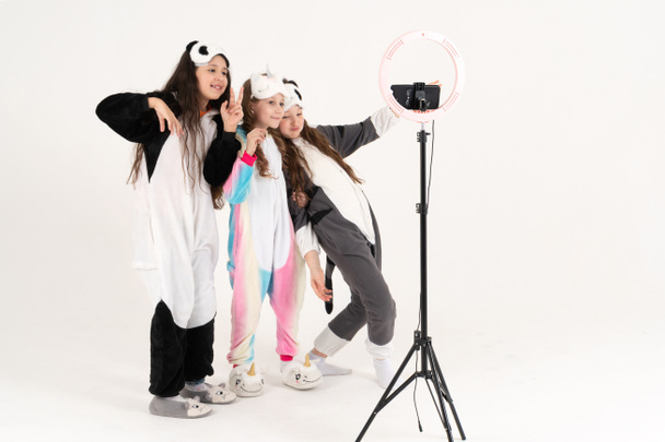 Χαριτωμένα έφηβα κορίτσια σε kigurumi και μάσκες ύπνου χαμογελώντας και πυροβολεί ένα βίντεο. Σέλφι. Το τηλέφωνο είναι τοποθετημένο σε ένα τρίποδο και η λάμπα δαχτυλίδι λάμπει - Φωτογραφία, εικόνα
