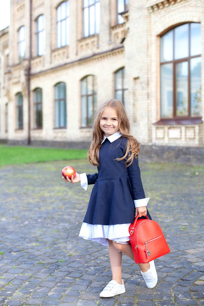 μαθήτρια του δημοτικού σχολείου με κόκκινο σακίδιο και μήλο στο δρόμο για τη μελέτη. Κοριτσάκι με φόρεμα και μακριά μαλλιά έξω κοντά στο κτίριο του σχολείου. Πίσω στο σχολείο. Εκπαιδευτική ιδέα. Το παιδί πάει για μάθημα - Φωτογραφία, εικόνα