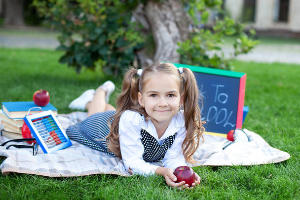 Wracać do szkoły. Cute uśmiechnięta uczennica siedzi na trawie z jabłkiem, książki w pobliżu szkoły. Edukacja przedszkolna. skopiować przestrzeń. Mała dziewczynka podczas lunchu uczy się na trawniku w parku. Koncepcja edukacji.  - Zdjęcie, obraz