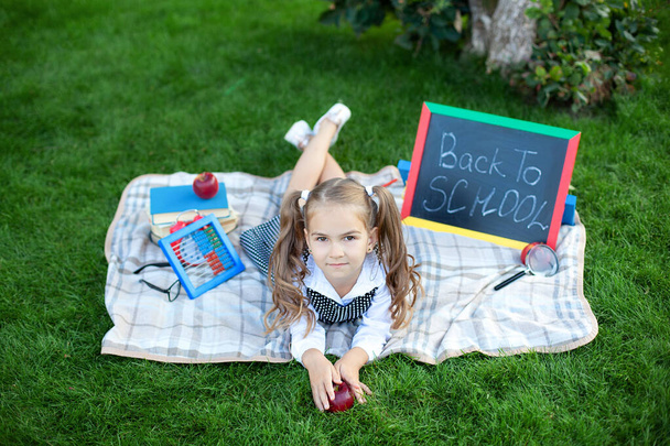 Мила усміхнена школярка сидить на траві з обідом, книжки біля школи. Повернутися до школи. Концепція освіти. Дошкільна освіта. копіювання простору. маленька дівчинка на обід і вчиться на газоні в парку
.  - Фото, зображення