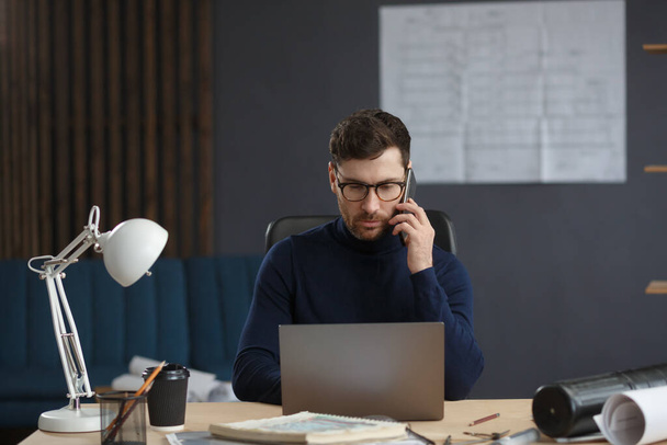 Architect talking on the phone in office. Confident businessman negotiates with business partners. Business portrait of handsome bearded man wearing eyeglasses sitting at workplace. Business concept - 写真・画像