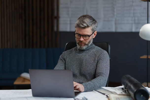 Designer working in office with laptop. Architect thinks over architectural plan, searching new ideas for construction project. Portrait of handsome bearded man sitting at workplace. Business concept - Foto, Imagen