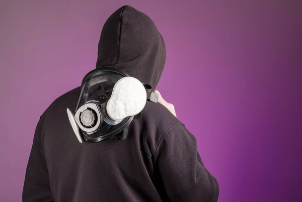 un hombre en una capucha con una máscara de gas en el hombro derecho nos dio la espalda. fondo púrpura - Foto, Imagen