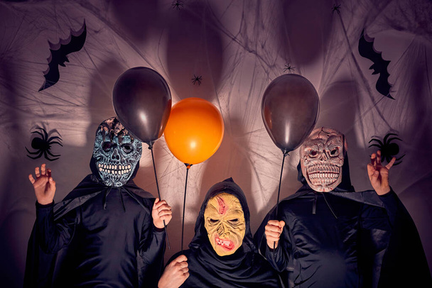 Zwei 7-jährige Jungen mit ihrer Mutter in Halloween-Kostümen mit Masken als Monster und einer Hexe mit schwarzen und orangefarbenen Luftballons. Dunkler Hintergrund mit Schatten, Spinnen, Spinnweben und Fledermäusen. - Foto, Bild