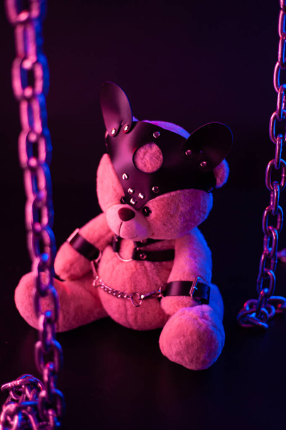 speelgoedbeer gekleed in lederen riemen harnas accessoire voor BDSM games op een donkere achtergrond in neon licht - Foto, afbeelding