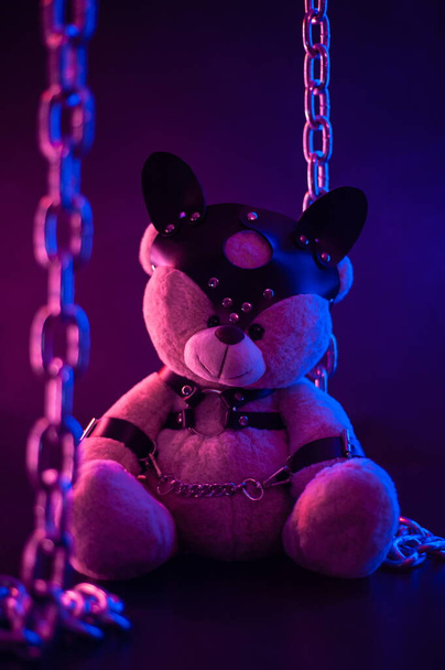 игрушечный медведь, одетый в кожаные ремни безопасности аксессуар для БДСМ игр на темном фоне в неоновом свете - Фото, изображение
