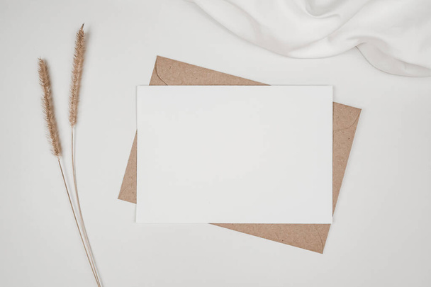 Prázdný bílý papír na hnědé papírové obálce s kvítkem a bílou látkou. Posunutí vodorovného prázdného pozdravného lístku. Horní pohled na obálku Craft na bílém pozadí. Plochý minimalismus - Fotografie, Obrázek