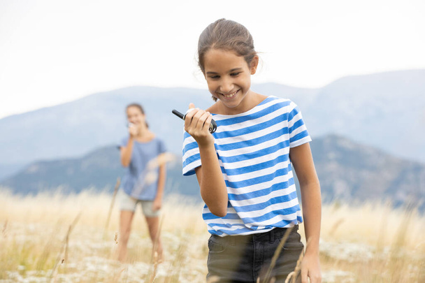Chica hablando en el walkie-talkie mientras su hermana la escucha en el fondo durante un paseo familiar en el prado - Foto, imagen