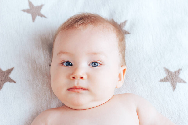 Egy fotó egy érzelemmentes babáról, egy újszülött kék szemekkel és szőke hajjal, amint egy világos takarón fekszik barna csillagokkal.. - Fotó, kép