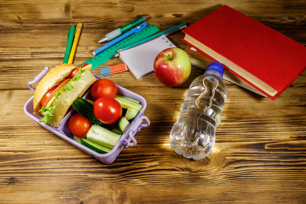 De vuelta al concepto escolar. Material escolar, libros, botella de agua, manzana y lonchera con hamburguesas y verduras frescas sobre una mesa de madera - Foto, Imagen