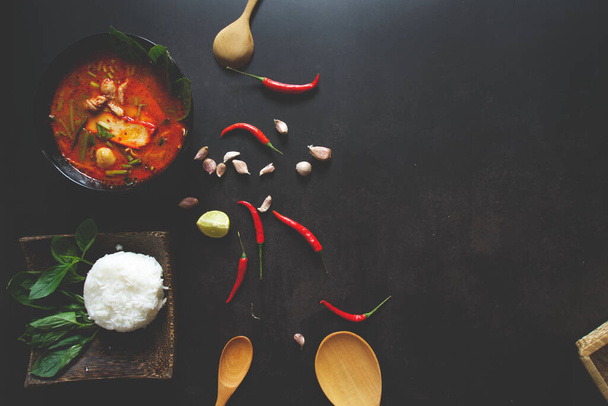 Thai food background concept. Dish of Thailand cuisine. Tom yum soup in black dish, rice in wooden plate, wooden ladle and spoon, chilis, garlic, lemon on table dark background with space for text at right - Photo, Image