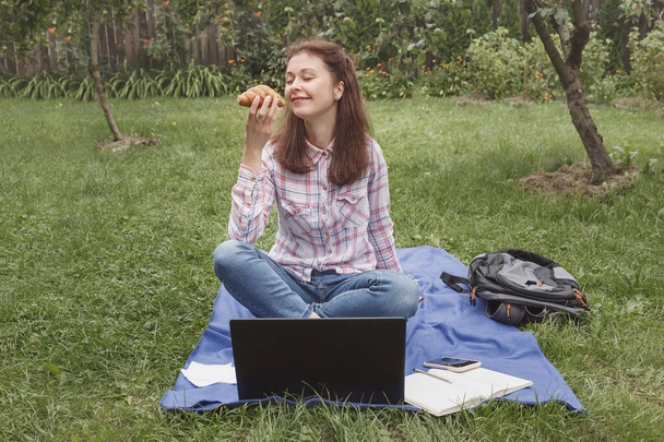 Młoda kobieta z rogalikiem w ręku siedzi w ogrodzie na trawie w pobliżu laptopa - Zdjęcie, obraz