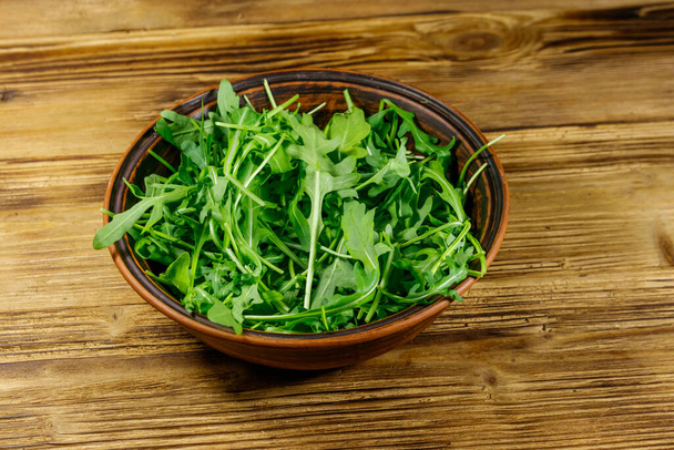 Свежая зеленая руккола в керамической чаше на деревянном столе. Здоровое питание или вегетарианская концепция - Фото, изображение