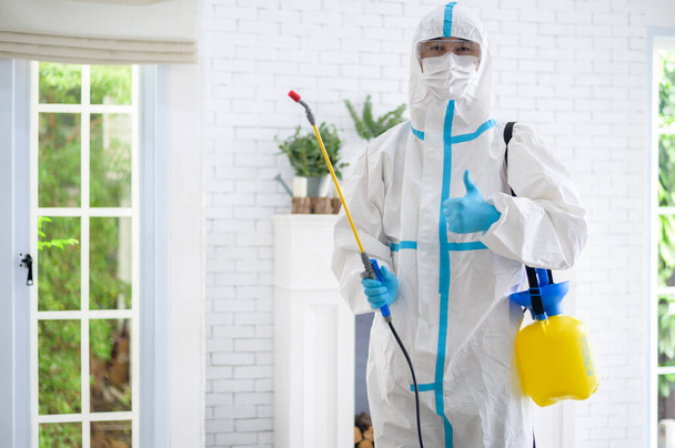 Lékařský personál v obleku PPE používá dezinfekční sprej v obývacím pokoji, Covid-19 ochrana, dezinfekce koncepce . - Fotografie, Obrázek
