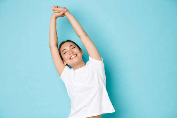 Portrait of carefree and relaxed, beautiful asian girl close eyes and stretching hands upwards with happy smile, feeling fresh and cheerful after taking nap, standing blue background - 写真・画像