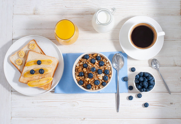 Breakfast with granola and blueberry, coffee, milk with orange juice and baked toasts on the white wooden table flat lay with close-up. Healthy tasty food at the morning with oat-flakes - Photo, image