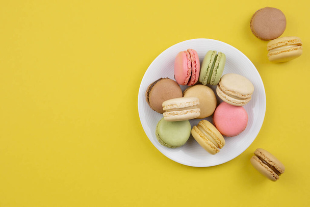 French colorful macaroons on a white plate. Yellow background. Top view. Copy space. - Фото, зображення