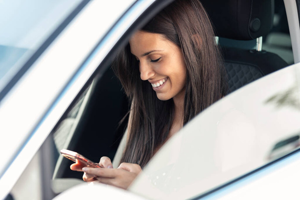 Portrait of beautiful young woman sending messages with her mobile phone in the car. - Фото, изображение