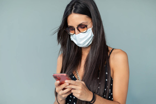 Portrait of beautiful young woman in face mask sending messages with her mobile phone while standing in the street. - Photo, image