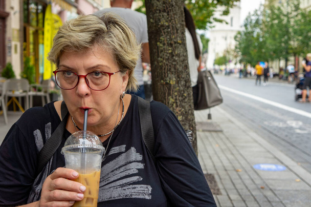 Una mujer madura y triste con gafas camina por las calles de la ciudad vieja. Bebe un cóctel en una mesa de café de la calle y mira a la distancia. - Foto, Imagen