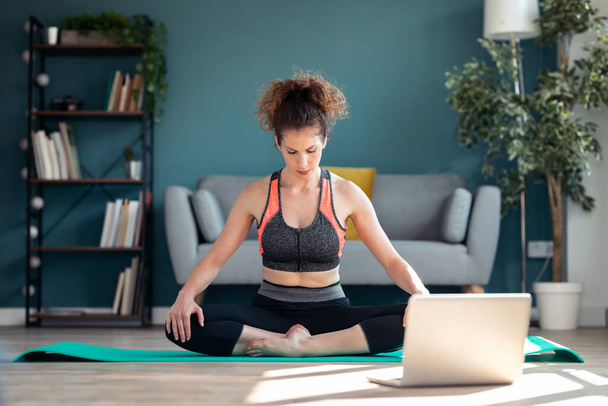 Shot of confident sporty young woman doing hypopressive exercises following online gym classes via laptop on floor in her living room at home. - Фото, изображение