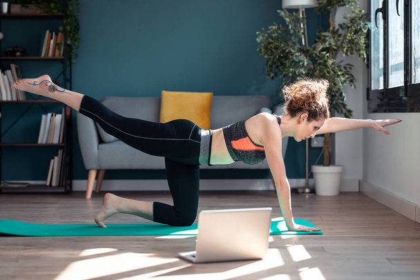 Shot of confident sporty young woman doing hypopressive exercises following online gym classes via laptop on floor in her living room at home. - Zdjęcie, obraz