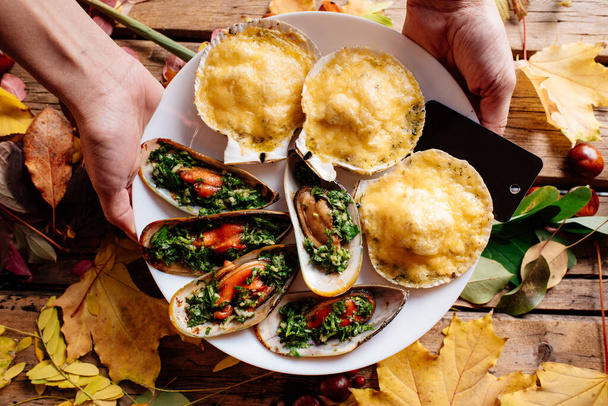 Baked mussels and scallops baked under cheese on white plate in hands of woman on wooden table with autumn leaves and chestnuts. - Foto, imagen