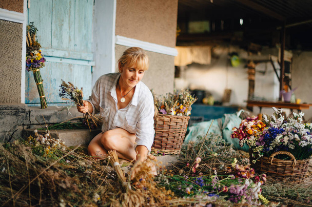 Positive woman in a shirt at the cottage creates bouquets of dried flowers with a smile on his face. Amateur florist makes compositions from flowers in the country. - Фото, изображение