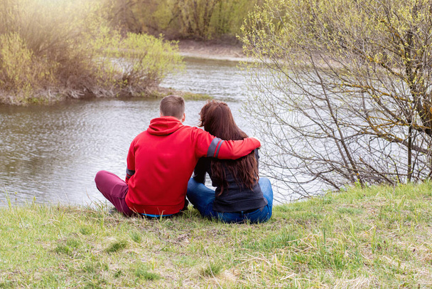 Νεαρό ζευγάρι αγκαλιάζει κάθεται στην όχθη του ποταμού. - Φωτογραφία, εικόνα