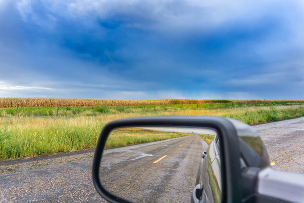 Zon op maïsvelden onder donkerblauwe wolken in Texas vooruit en in achteraanzicht spiegel de weg achter ons. - Foto, afbeelding