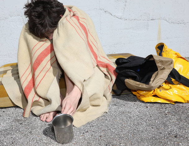 fiatal hajléktalan férfi ül egy matracon, és a koszos takaró alatt pénzt kér az emberektől a városban. - Fotó, kép