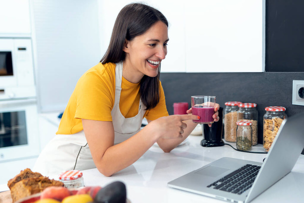 Shot of beautiful young nutritionist woman having an online video call via laptop computer with a friend to showing detox handmade smoothie in the kitchen at home. - Φωτογραφία, εικόνα