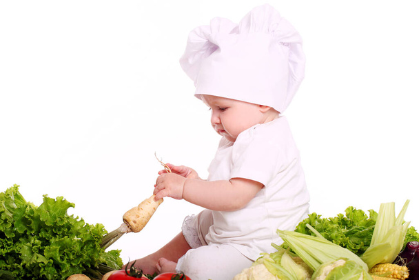 Bonito chef bebê com diferentes vegetais sobre fundo branco
 - Foto, Imagem