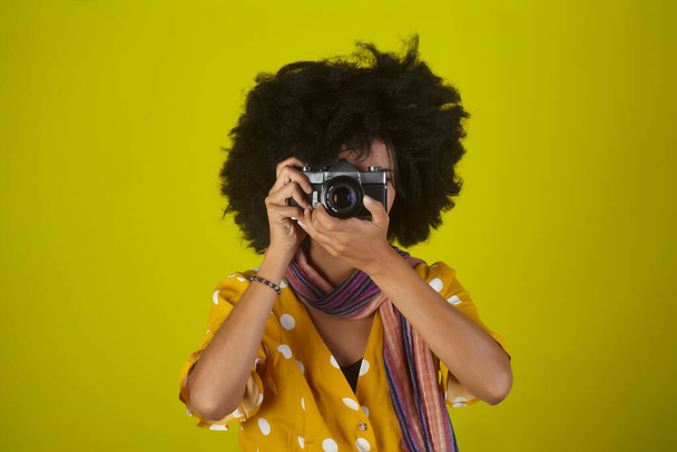 Mooi indisch meisje met afro krullend kapsel praten foto 's met retro camera op gele achtergrond - Foto, afbeelding
