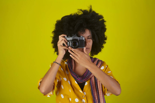 Beautiful indian girl with afro curly hairstyle talking pictures  with retro camera on yellow background - Foto, imagen
