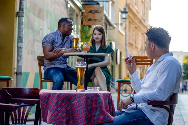  Multiracial couple on a date at a street terrace of the cafe. Third man smoking cigarette and looking at the woman from distance. Concept of diversity and jealousy. Selective focus. - Photo, image