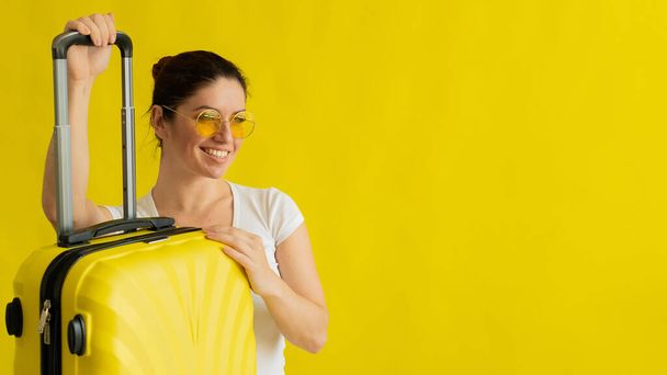 Lächelnde Frau mit Sonnenbrille und Koffer mit ausklappbarem Griff auf gelbem Hintergrund - Foto, Bild