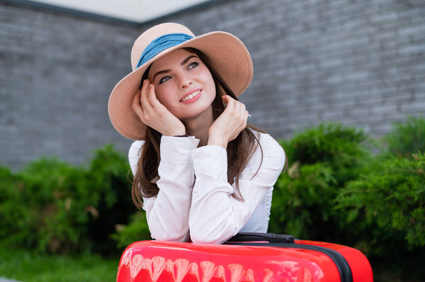 Portrait of a happy beautiful caucasian woman in a hat with a big red suitcase outdoors. Attractive girl leaned against luggage while waiting for a taxi. - Foto, Bild