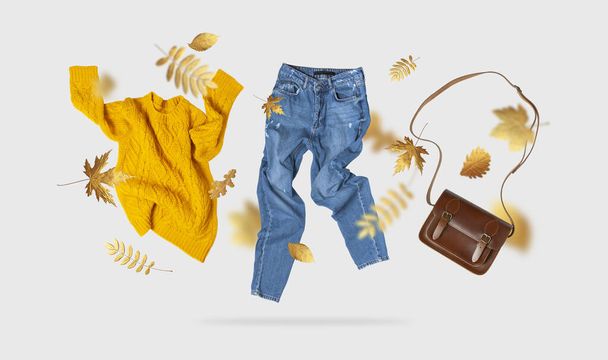 Yellow flying women's knitted sweater blue jeans brown leather bag golden autumn leaves on gray background. Creative clothing concept, trendy fall cozy sweater pullover jersey. Female flat lay fashion. - Photo, Image