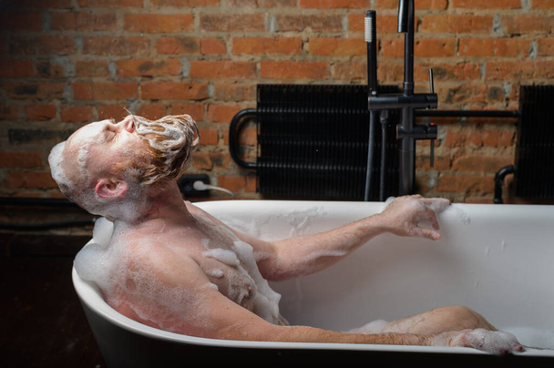 Close-up portrait of funny red-bearded bald man takes a bath with foam on the background of a brick wall. Cheerful naughty guy at spa treatments. A parody of glamorous girls. - Foto, afbeelding