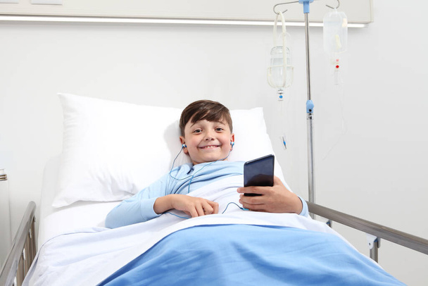 bambino felice in letto d'ospedale utilizzando smartphone naviga su internet indossando auricolari - Foto, immagini