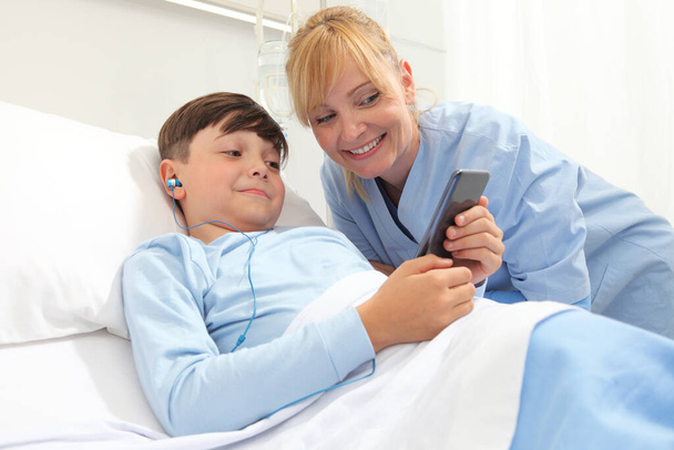 Szczęśliwe dziecko leżące w łóżku w pokoju szpitalnym i uśmiechnięta pielęgniarka korzystająca ze smartfona surfuje po Internecie nosząc słuchawki - Zdjęcie, obraz