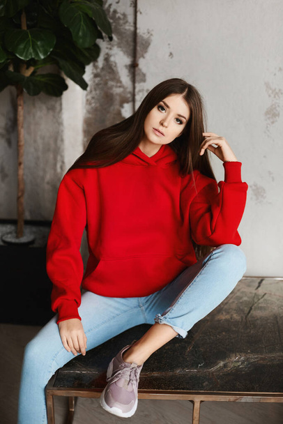 Young hipster girl in a red hoodie and blue jeans posing in the loft interior - Photo, image