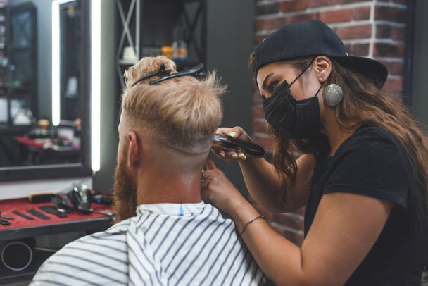 Peluquero mujer con máscara corta los pelos de un hombre con cortador de pelo. peinado durante el distanciamiento social. - Foto, imagen