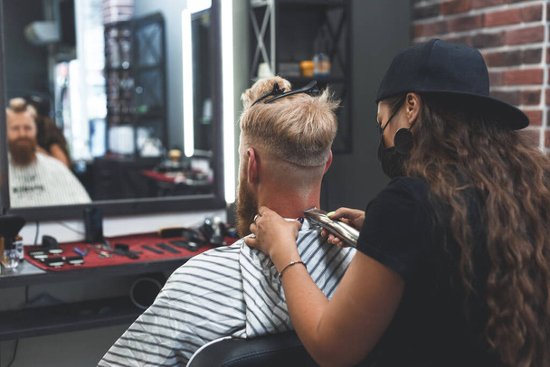 Eine Friseurin in Maske schneidet einem Mann mit Haarschneidemaschine die Haare. Frisur bei sozialer Distanzierung. - Foto, Bild